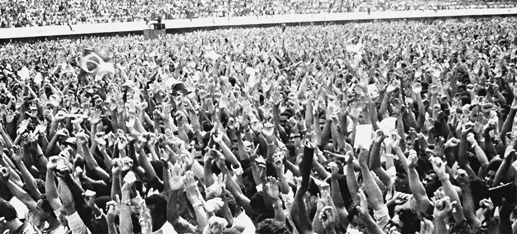 Fotografia em preto e branco. Multidão reunida. Todos estão com os braços levantados.