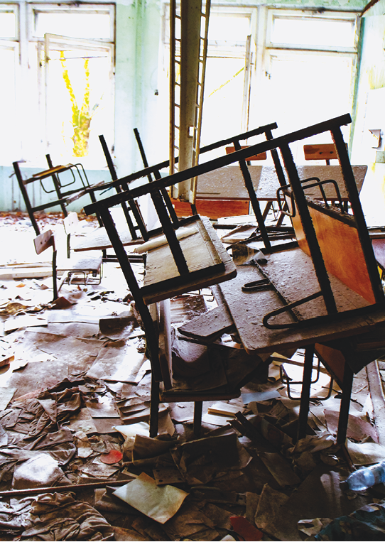 Fotografia. Vista do interior de uma sala de aula em ruínas, com carteiras e móveis quebrados e amontoados e uma camada de lixo e de escombros encobrindo o chão.