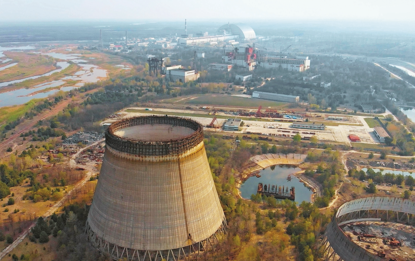 Fotografia aérea. Vista da Usina nuclear de Chernobyl. À frente, grande construção cilíndrica. Ao redor, vegetação. À direita, um tanque de água. Ao fundo, edifícios da usina e, à esquerda, um rio parcialmente seco.