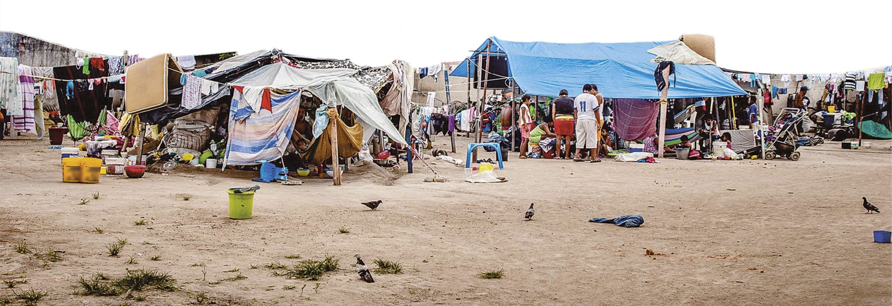 Fotografia. Pessoas em um acampamento improvisado em uma área de terra batida. As tendas são feitas com lonas e tecidos sobrepostos, presos em estacas. Os pertences das pessoas estão aglomerados sob as tendas. À esquerda, há um varal pendurado fora de uma tenda, com roupas de crianças, roupas de cama e um colchão pendurado.