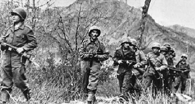 Fotografia em preto e branco. Soldados armados em fila andando entre o mato alto.