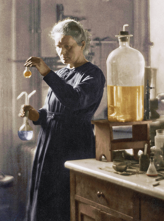 Fotografia. Marie Curie, senhora de vestido escuro em um laboratório. Ela manuseia dois frascos de vidro. Ao seu lado, uma bancada com outros frascos e equipamentos.