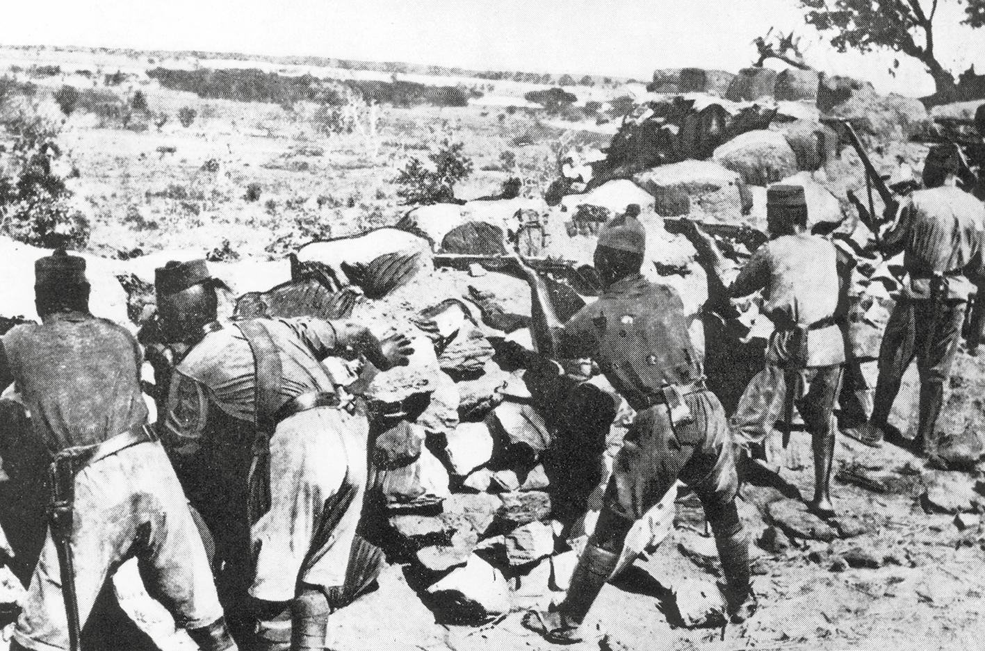 Fotografia em preto e branco. Soldados atrás de uma trincheira feita de pedras. Empunham armas de cano longo sobre a trincheira.