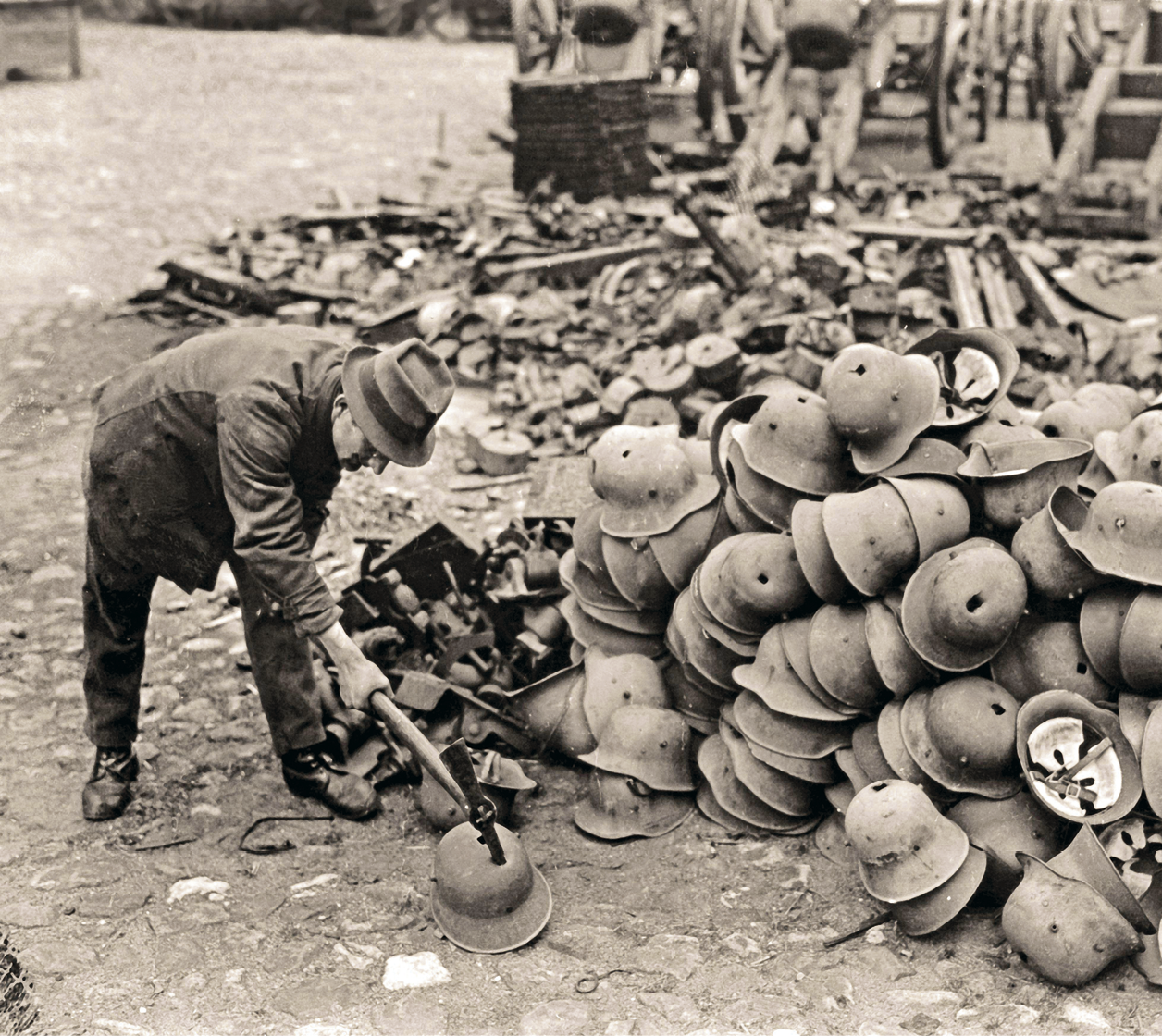 Fotografia em preto e branco. Homem de terno e chapéu martelando capacetes. Alguns com um furo no centro estão empilhados à direita. Ao fundo, outros materiais aglomerados.