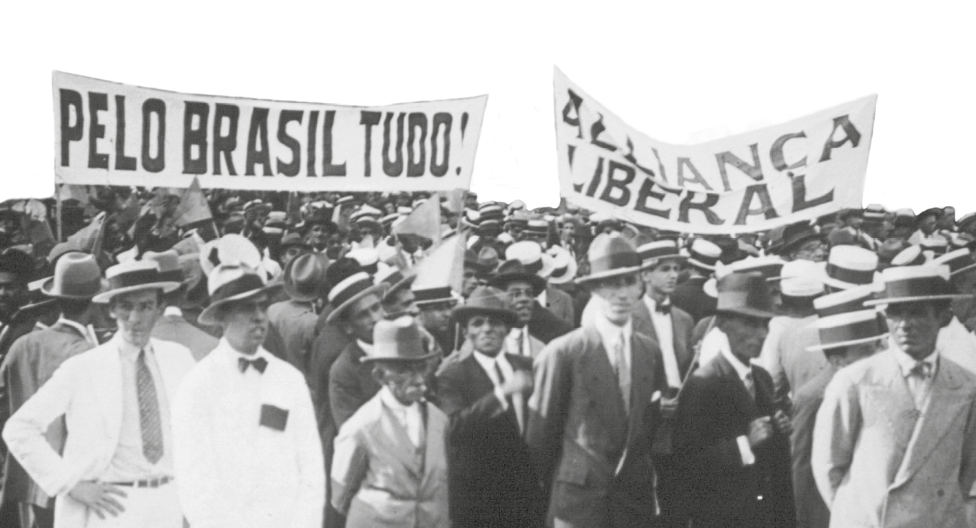 Fotografia em preto e branco. Homens de terno, gravata e chapéu, reunidos. Carregam faixas com os dizeres: PELO BRASIL TUDO! e ALIANÇA LIBERAL.