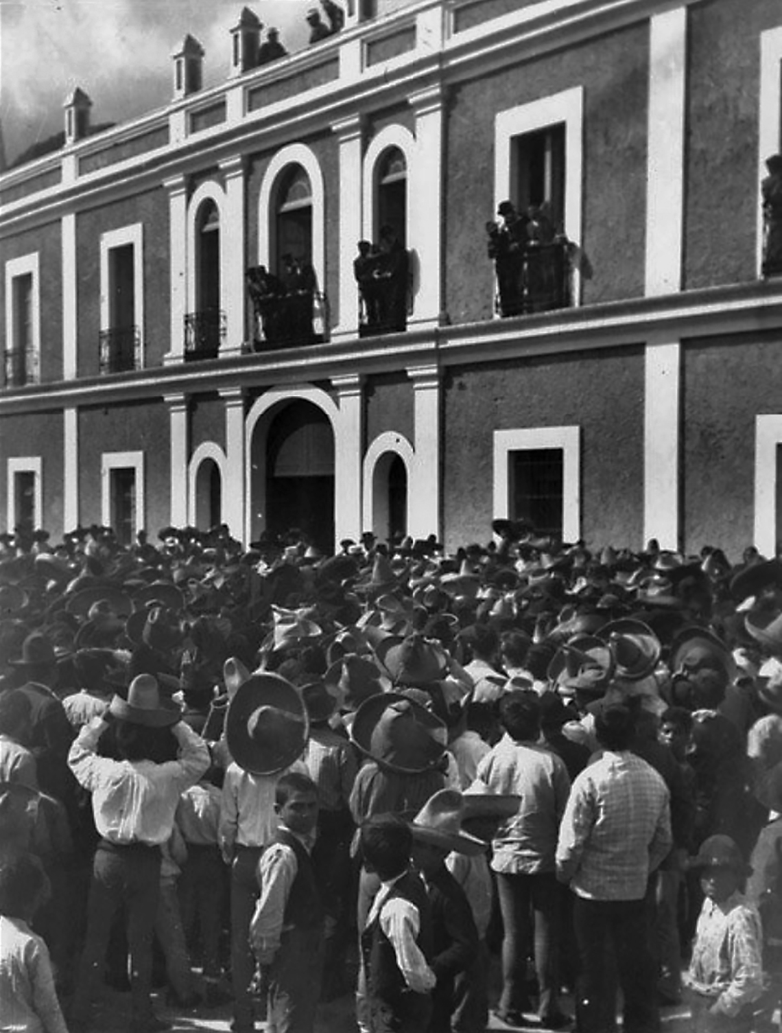 Fotografia em preto e branco. Pessoas reunidas em frente a um prédio. Eles observam alguns homens na sacada, no andar de cima.