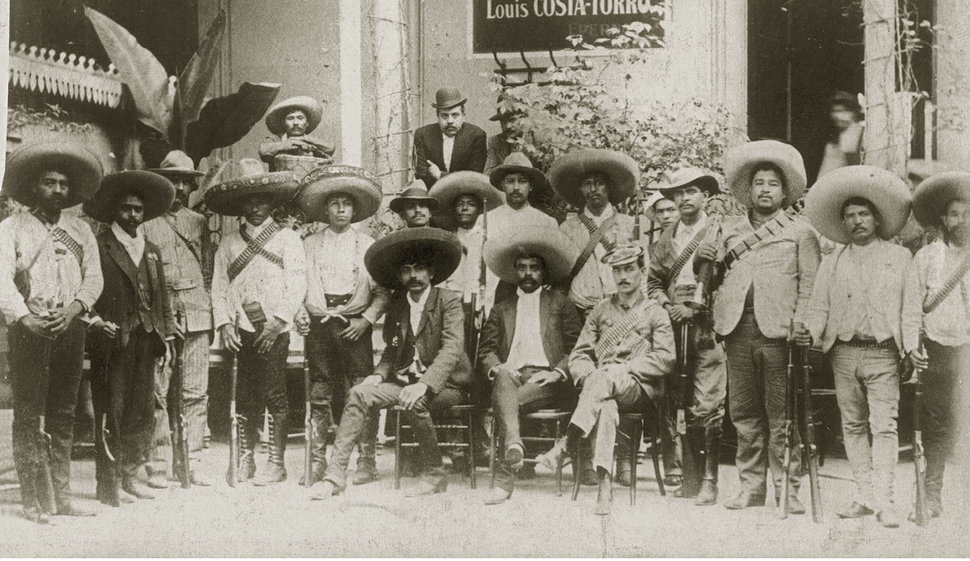 Fotografia em preto e branco. Emiliano Zapata ao centro, sentado de pernas cruzadas. Veste camisa clara e um terno escuro aberto. Usa um grande chapéu redondo. Sentados ao lado dele um militar e um homem de bigode e chapéu grande.  Ao redor deles, em pé, outros homens de chapéu. Alguns seguram armas.