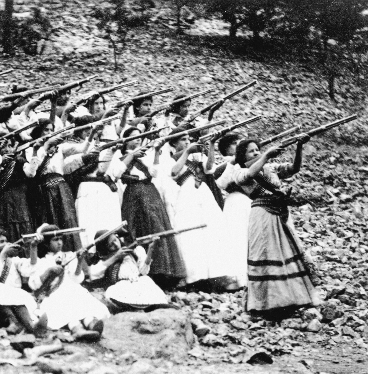 Fotografia em preto e branco. Grupo de mulheres na parte baixa de uma colina. Estão empunhando e apontando armas de cano longo para o alto, à direita.