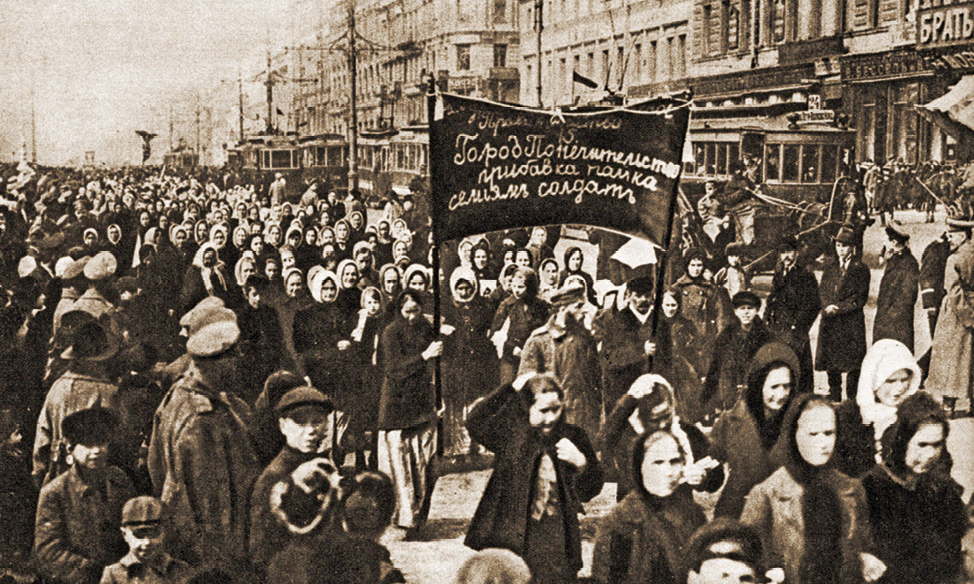 Fotografia em preto e branco. Pessoas na rua, em sua maioria mulheres, em um protesto. Usam roupas escuras e carregam uma faixa com dizeres em russo.