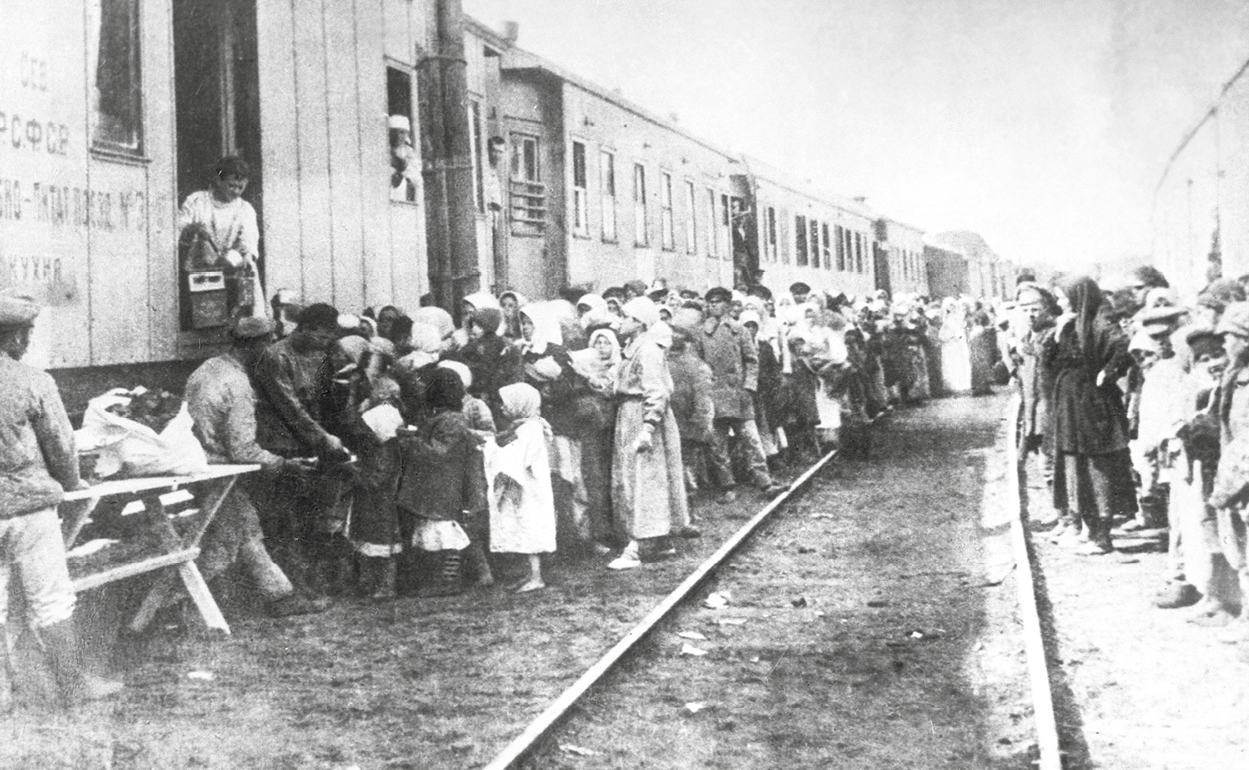 Fotografia em preto e branco. Pessoas em duas longas filas ao redor de um trem. À esquerda, elas recebem comida de uma pessoa em um dos vagões do trem. No centro da imagem, trilhos.