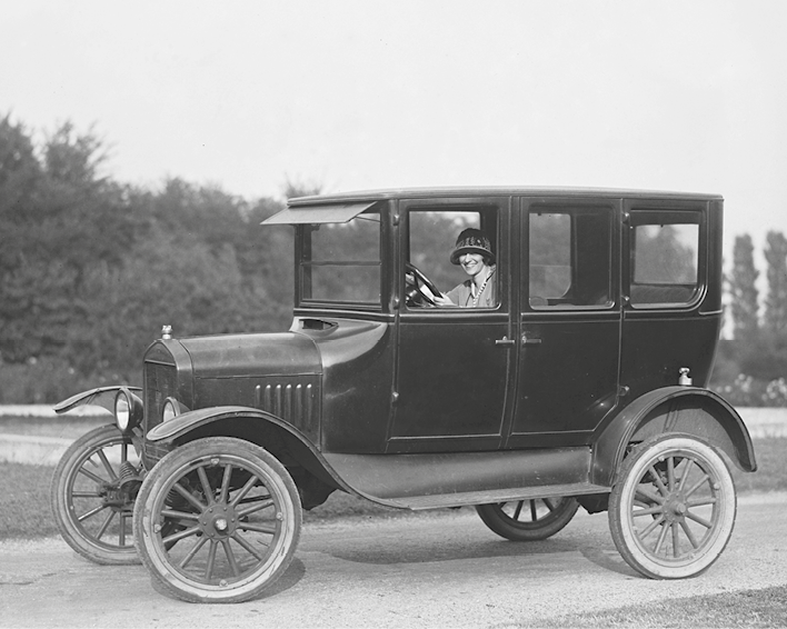 Fotografia em preto e branco. Mulher sorridente de chapéu dirigindo um veículo com quatro rodas finas, estrutura alta e quadrada. Tem janelas e portas dianteiras e traseiras.