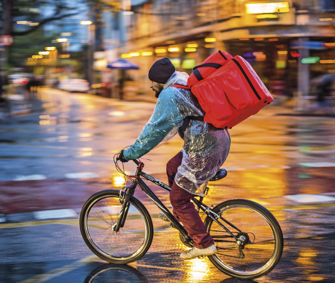 Fotografia. Entregador de comida em bicicleta. Ele carrega uma mochila grande e vermelha às costas que contém um isopor. O entregador usa touca e capa de chuva. O asfalto está molhado. Ao fundo as luzes da rua desfocadas.