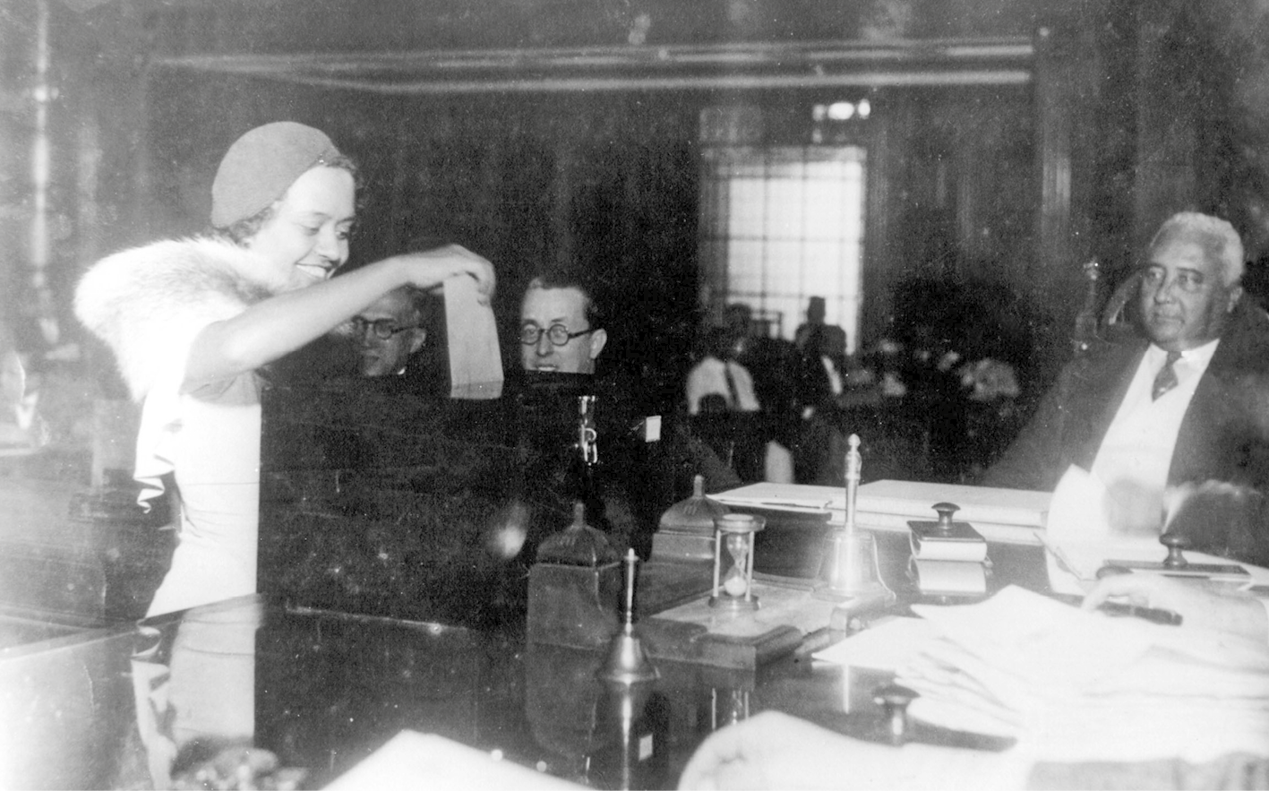Fotografia em preto e branco. Mulher sorridente, de vestido claro e chapéu, coloca um pedaço de papel dentro de uma urna. Homens sentados observam a cena.