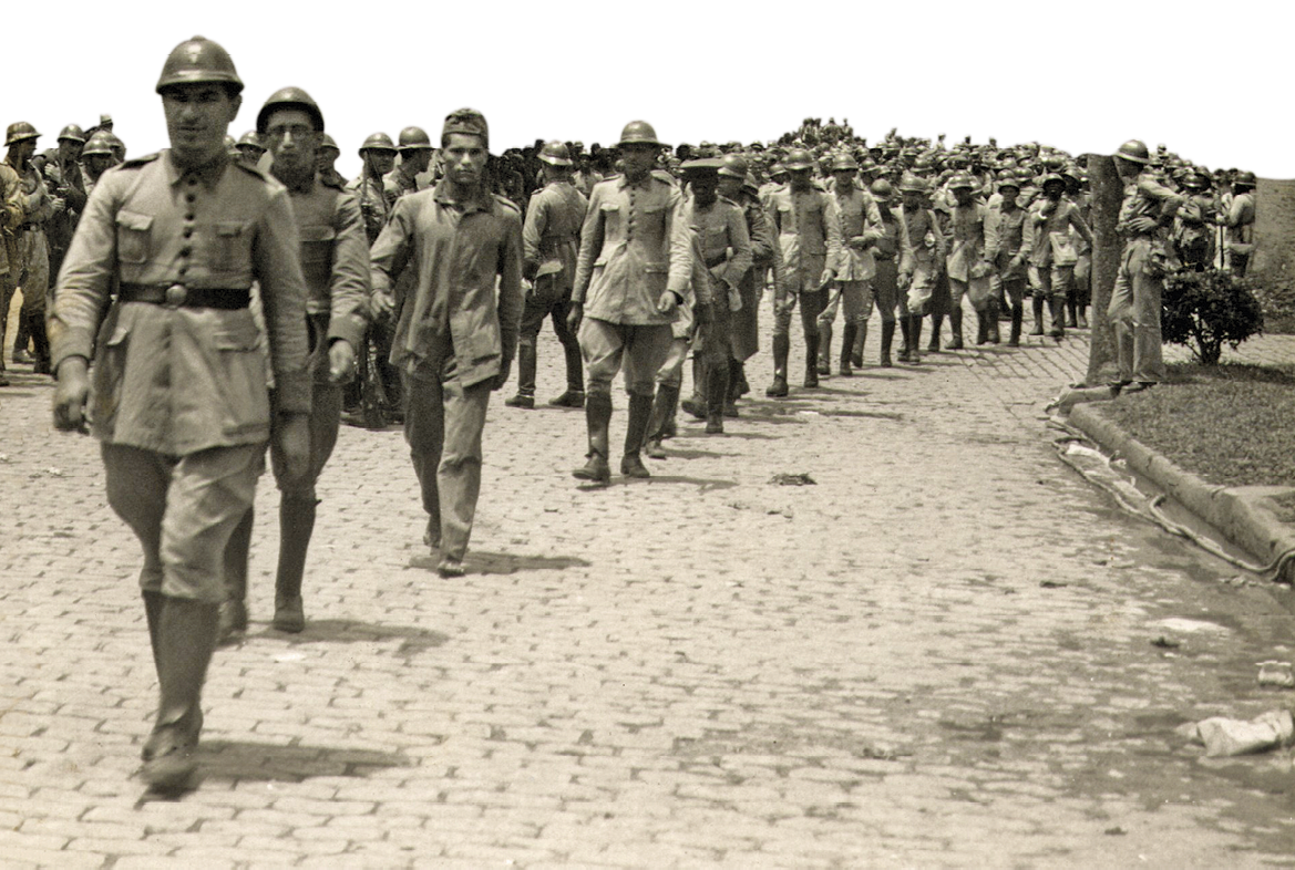 Fotografia em sépia. Homens de casaco largo em filas. Alguns usam fardamento militar e capacete.