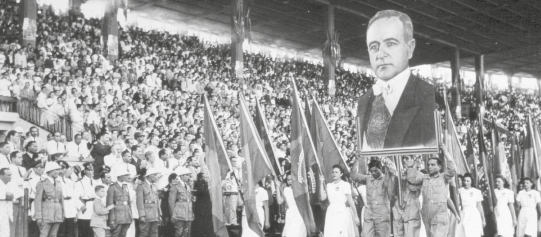 Fotografia em preto e branco. Multidão nas arquibancadas de um estádio. No centro dele, um grupo desfila com bandeiras e a imagem de Getúlio Vargas.