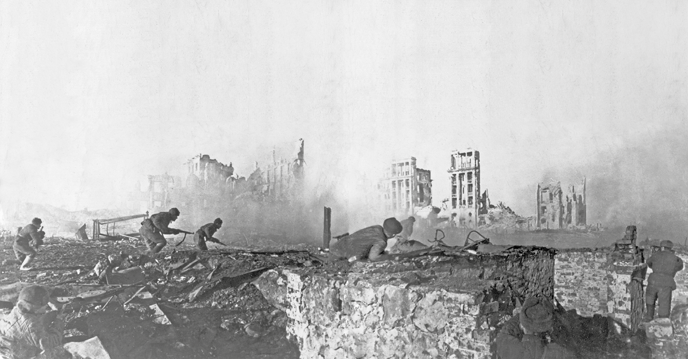 Fotografia em preto e branco. Soldados abaixados entre os escombros. Ao fundo muita fumaça e prédios destruídos.