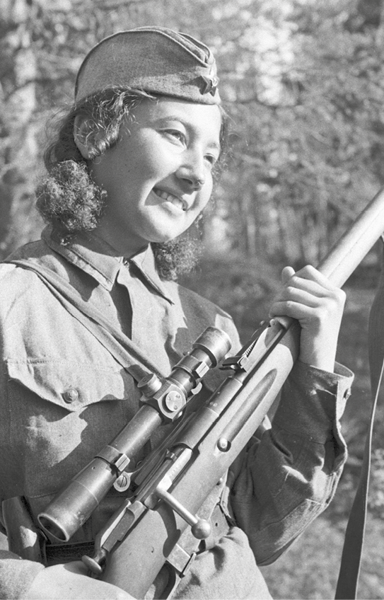 Fotografia em preto e branco. Mulher de cabelo cacheado, curto e rosto arredondado. Está sorrindo. Usa uma boina e segura uma arma de cano longo.