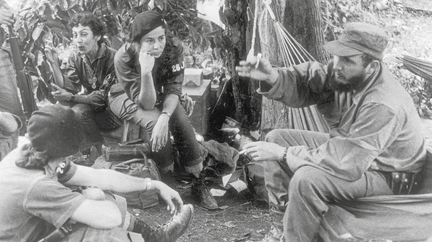 Fotografia em preto e branco. Fidel Castro, homem de boina, barba comprida e macacão. Está à direita, sentado em uma rede. À sua frente, algumas mulheres de macacão e boina sentadas no chão.