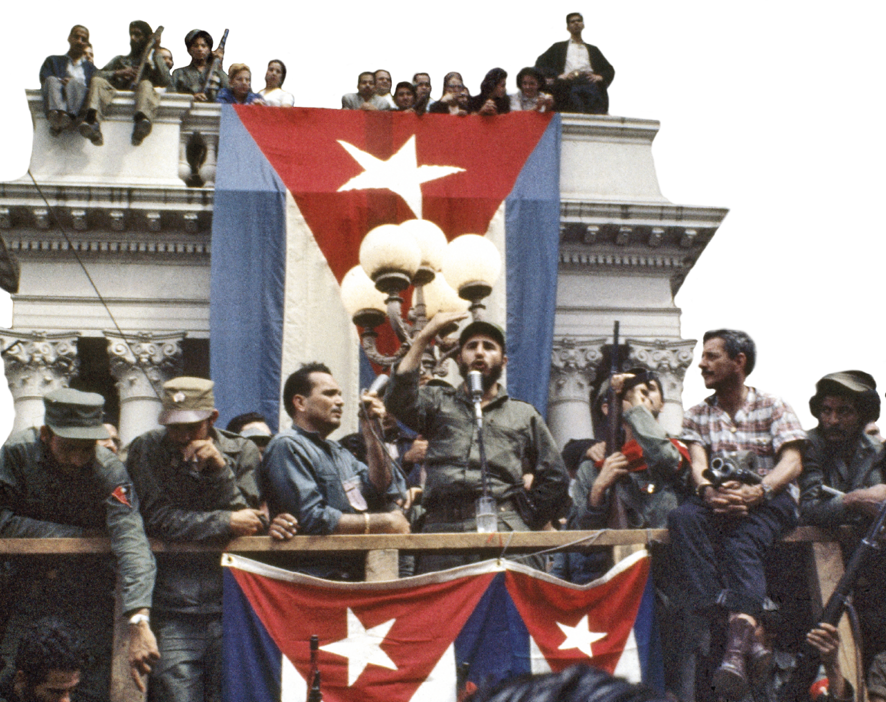 Fotografia. No primeiro plano, Fidel Castro sobre um palanque. Veste macacão verde e boina. Discursa em um microfone com uma das mãos levantadas. Ao seu redor, diversas pessoas assistem. Algumas carregam armas de fogo. Ao fundo uma grande bandeira de Cuba pendurada em um terraço em que se vê algumas pessoas. À frente de Fidel, duas bandeiras de Cuba igualmente penduradas.