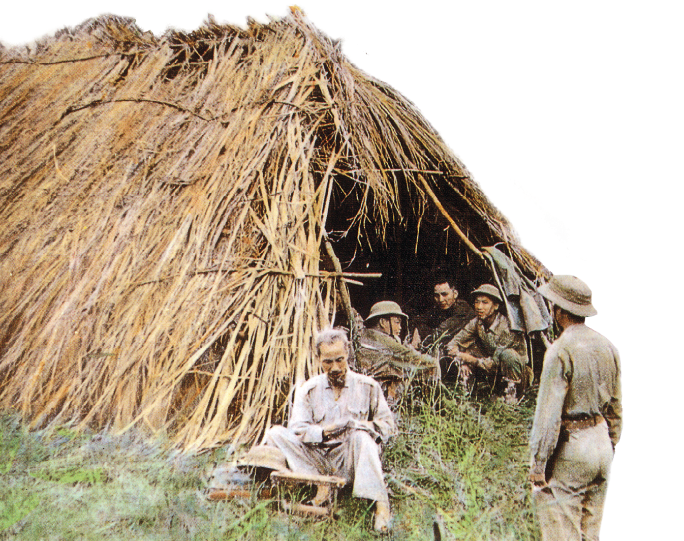 Fotografia. No primeiro plano, um senhor de cabelos brancos, roupas claras e largas está sentado na grama, à frente de uma cabana de palha. Dentro dela, alguns soldados.