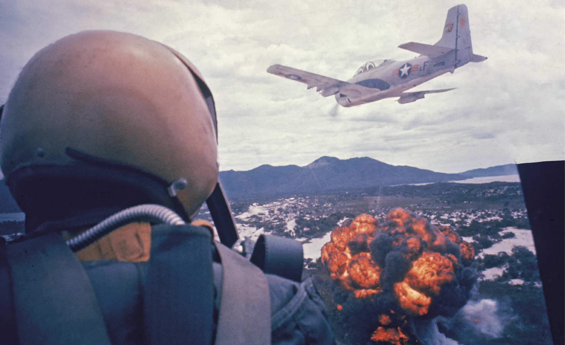 Fotografia. Em primeiro plano, piloto de costas, dentro de um avião militar. Ele está de capacete. Ao fundo, outro avião militar sobrevoa uma área em chamas.