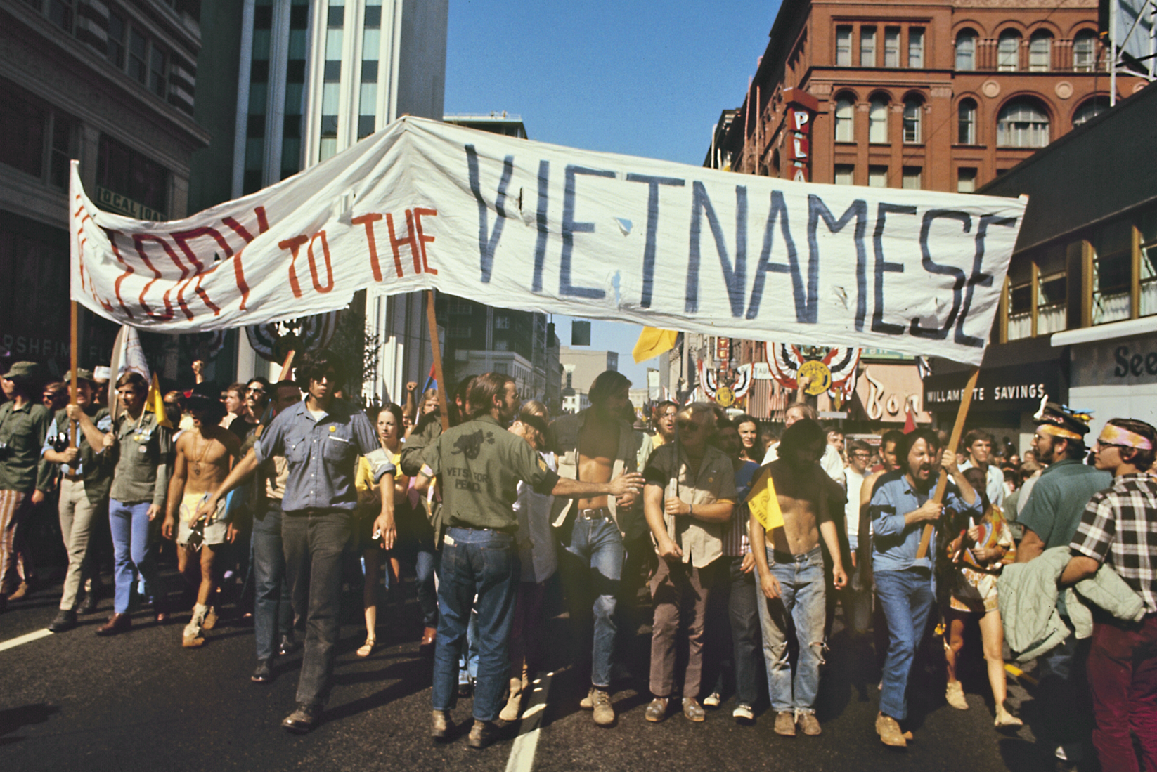 Fotografia. Pessoas na rua em um protesto. Elas carregam uma grande faixa com plavras de ordem em favor dos vietnamitas.