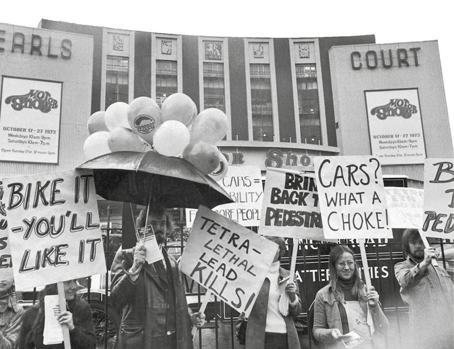 Fotografia em preto e branco. Algumas pessoas em um protesto na frente de um prédio. Elas carregam cartazes feitos à mão com palavras de ordem.