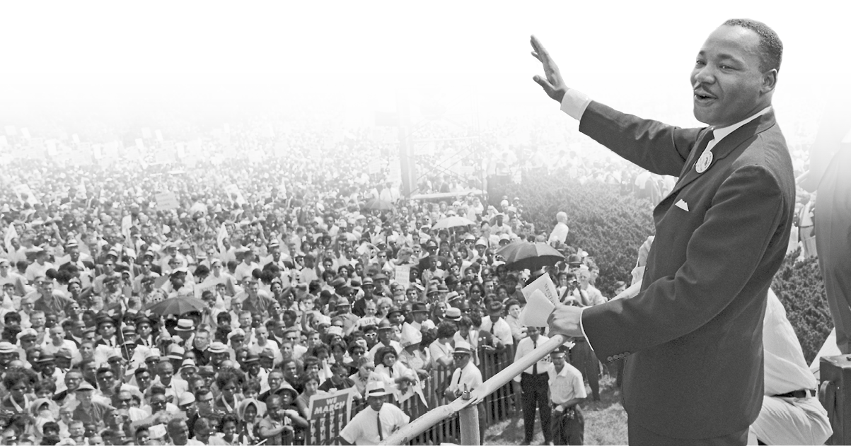 Fotografia em preto e branco. Martin Luther King, homem negro de rosto arredondado, cabelos curtos e bigode fino. Veste terno e gravata. Está sobre um palanque e acena para a multidão ao redor.
