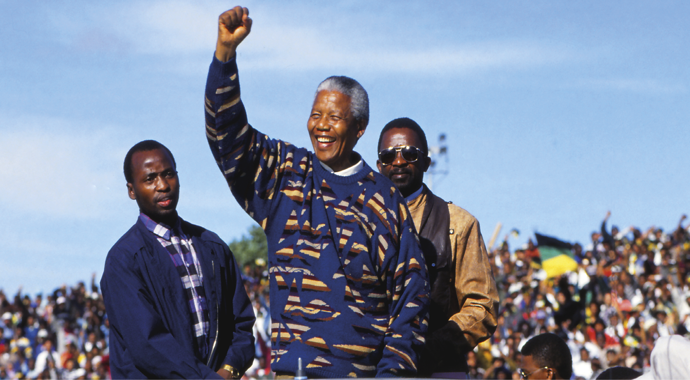 Fotografia. No primeiro plano, Nelson Mandela, senhor negro, sorridente. Saudando uma multidão com o braço direito estendido para cima e o punho cerrado. Atrás dele dois homens.
