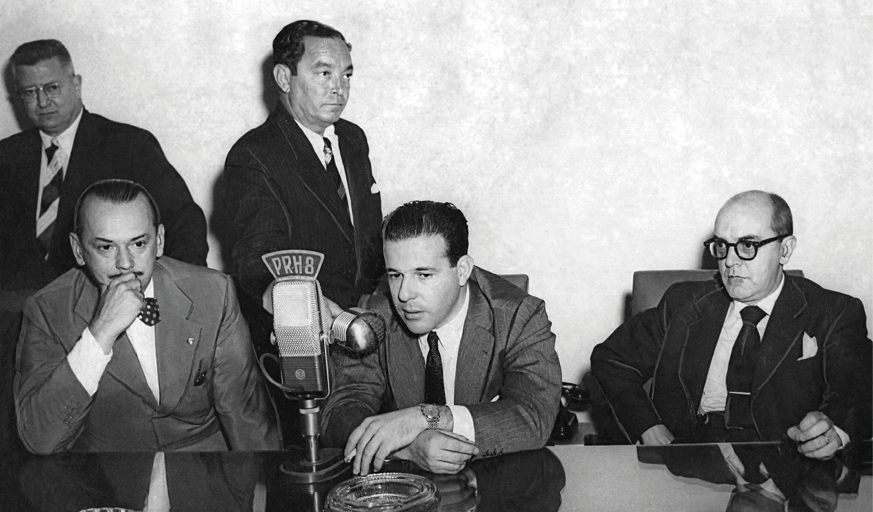 Fotografia em preto e branco. Três homens sentados à mesa. O homem do meio é Jango que está falando ao telefone.  Atrás deles, dois homens em pé.