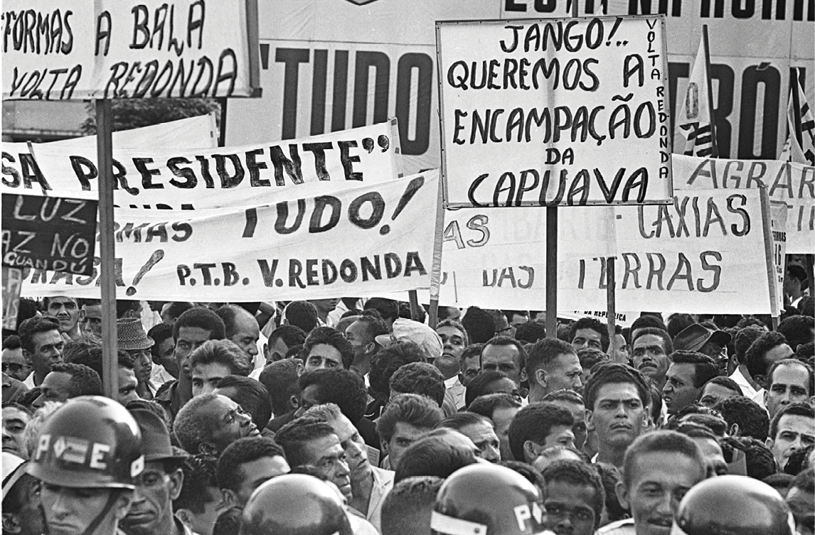 Fotografia em preto e branco. Pessoas aglomeradas carregando faixas e cartazes com os dizeres: JANGO QUEREMOS A ENCAMPAÇÃO DA CAPUAVA, dentre outros.