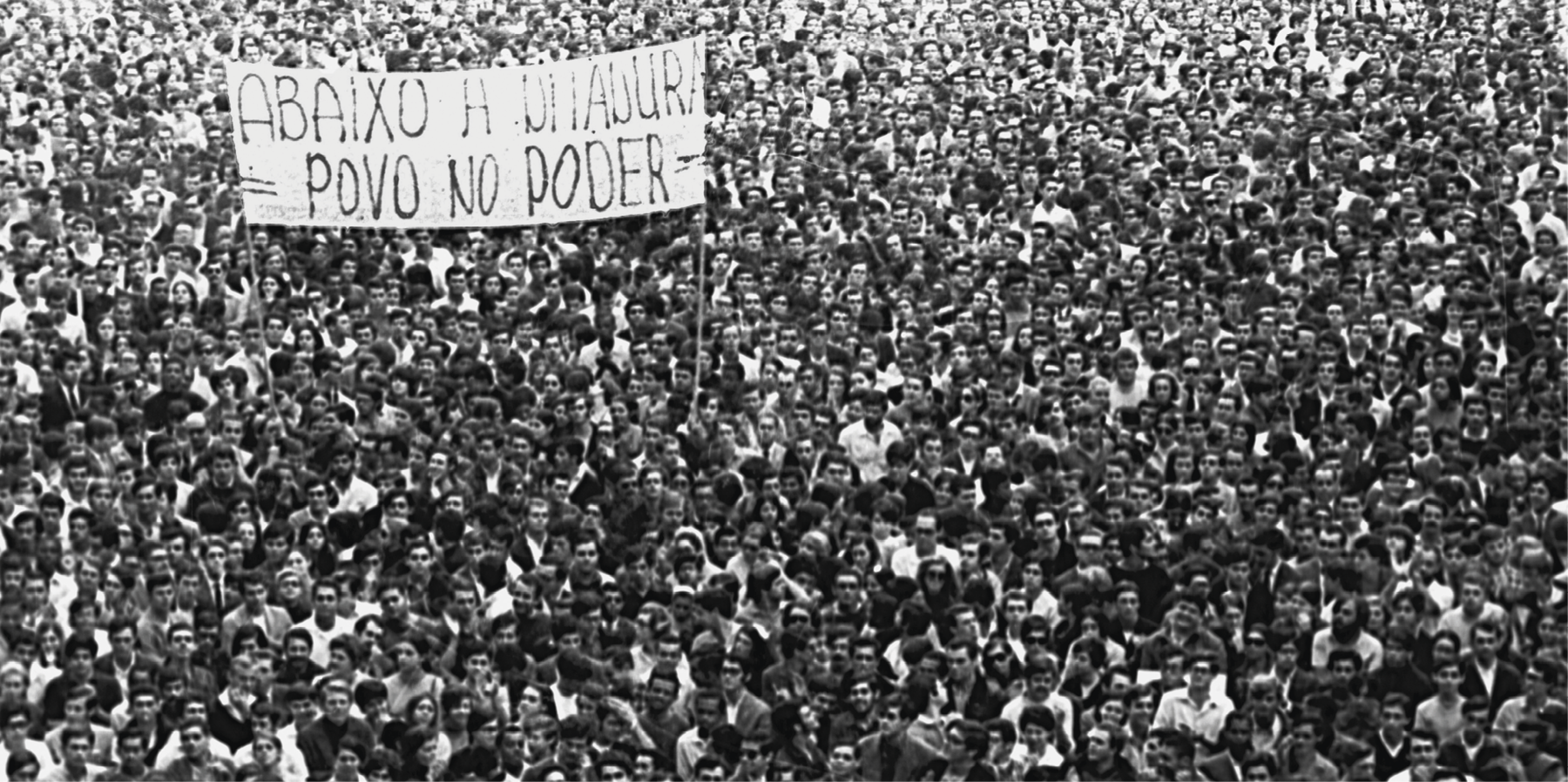 Fotografia em preto e branco. Multidão aglomerada em uma passeata. No centro, um grande cartaz com a frase: ABAIXO A DITADURA – POVO NO PODER.