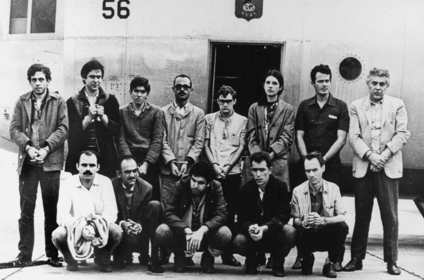 Fotografia em preto e branco. Treze pessoas de mãos algemadas, organizados em duas fileiras. As cinco pessoas na fileira da frente estão agachadas. Ao fundo, parte da fuselagem de um avião.