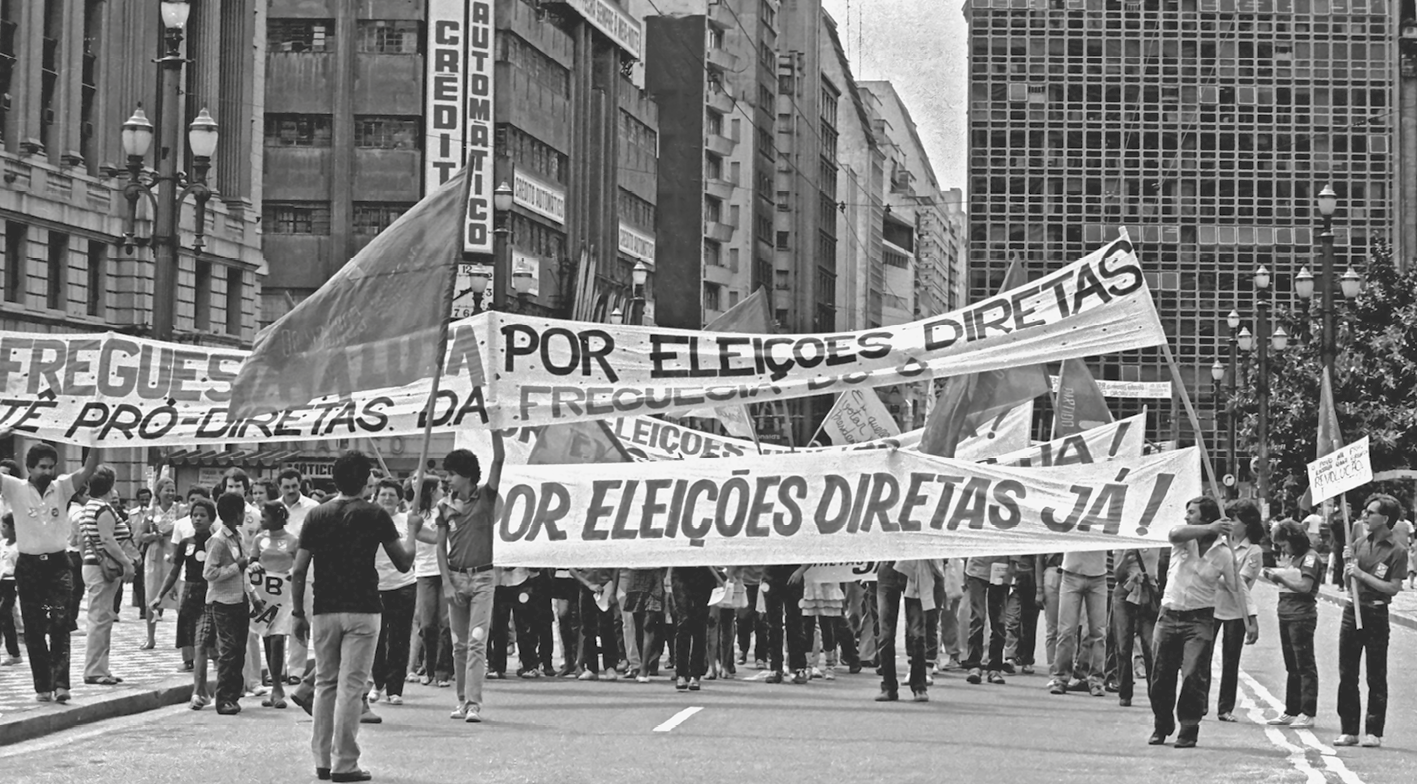 Fotografia em preto e branco. Pessoas na rua em uma manifestação. Carregam faixas com os dizeres: POR ELEIÇÕES DIRETAS JÁ!.