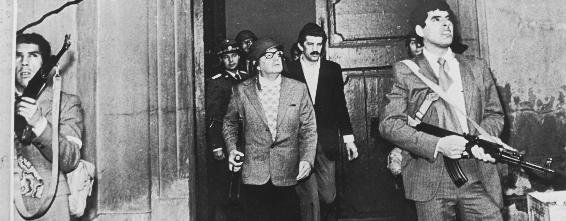 Fotografia em preto e branco. No centro da imagem, Salvador Allende, senhor de óculos e capacete. Está em frente a uma grande porta escoltado por outros homens. À sua frente, um homem com a testa franzida e os dentes cerrados. Empunha uma grande. Ao redor, outros homens.