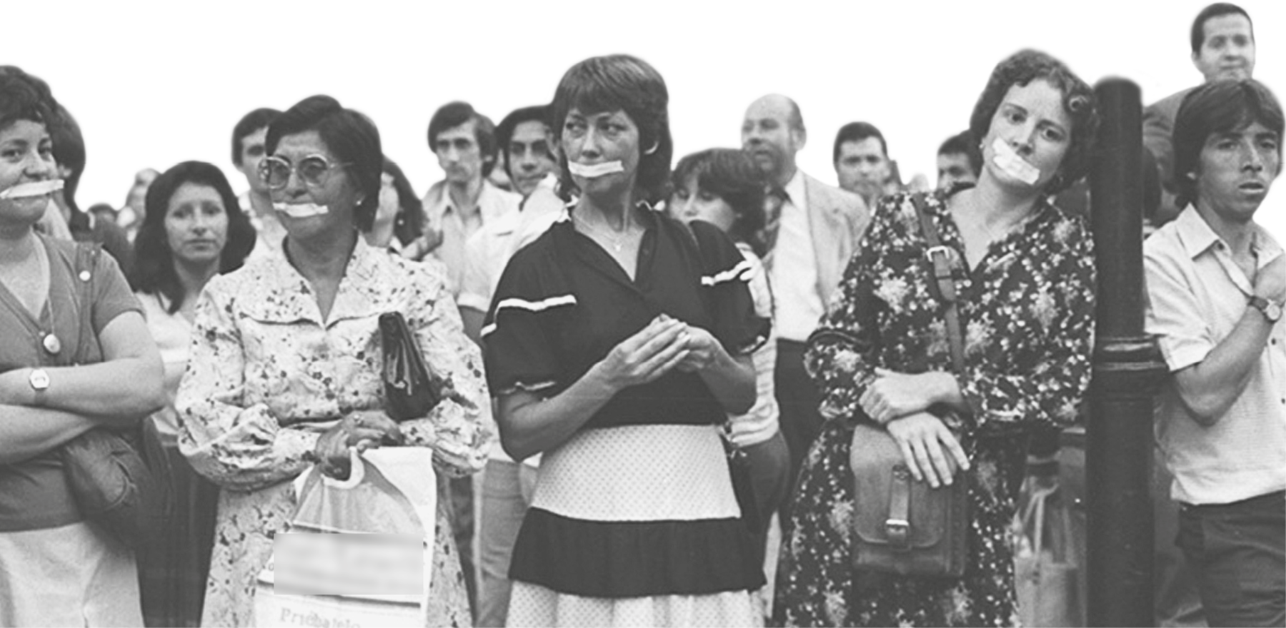 Fotografia em preto e branco. Grupo de pessoas reunidas em um protesto. As mulheres à frente têm uma fita sobre a boca.