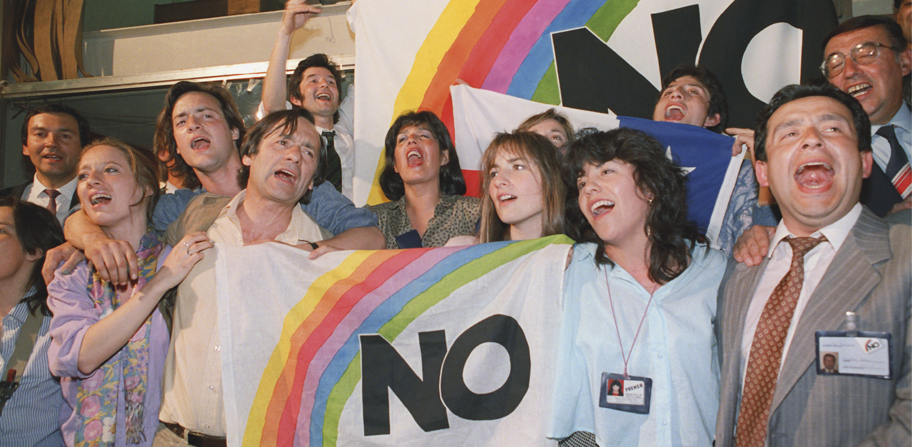 Fotografia. Grupo de homens e mulheres em uma celebração. Eles carregam uma bandeira branca com a palavra NO e um arco íris ao redor.