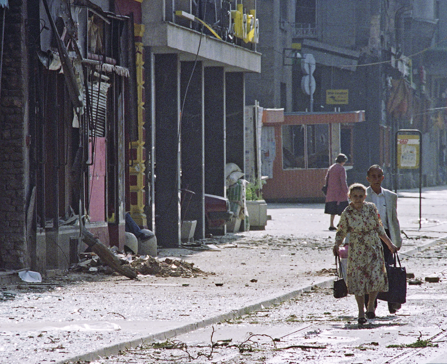 Fotografia. Três pessoas caminhando em uma rua. Os prédios estão parcialmente destruídos. No chão muito entulho, escombros e sujeira.