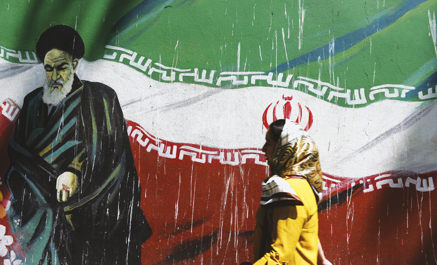 Fotografia. Ao fundo, um muro com uma pintura representando a bandeira iraniana: três listras horizontais, uma verde (acima), uma branca (no centro) e uma vermelha (abaixo), com inscrições islâmicas em branco acima e abaixo da faixa do centro e um emblema vermelho no centro da faixa branca. À esquerda, pintado diante da bandeira, um senhor de barba longa e branca, vestindo túnica e um turbante pretos. Em primeiro plano, à direita, uma moça com um lenço amarelo cobrindo os cabelos e um casaco amarelo passa diante do muro, caminhando da direita para a esquerda.