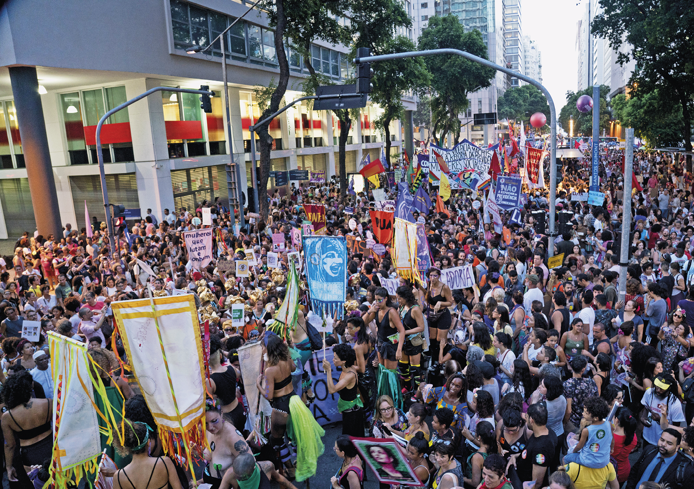 Fotografia. Vista do alto, uma multidão em uma manifestação em uma avenida larga. As pessoas carregam bandeiras, estandartes e faixas.  à esquerda, vista parcial de um prédio branco com janelas de vidro.