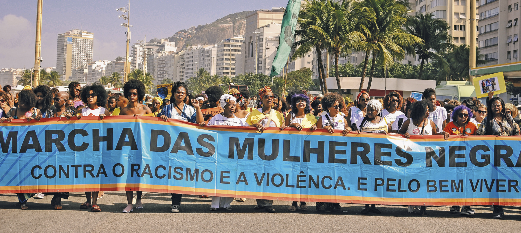 Fotografia. Mulheres em manifestação em uma rua. Elas carregam uma grande faixa de fundo azul com bordas cor de laranja com o texto: 'MARCHA DAS MULHERES NEGRAS CONTRA O RACISMO E A VIOLÊNCIA E PELO BEM VIVER'. Ao fundo, prédio, um morro e algumas palmeiras. No alto, o céu azul claro.
