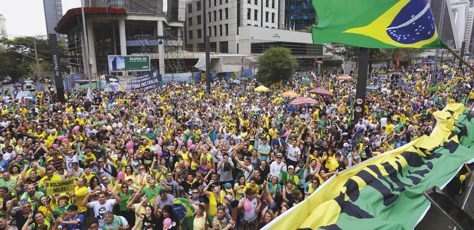 Fotografia. Pessoas vestidas com roupas nas cores verde e amarelo agrupadas em uma avenida em uma manifestação. À direita, uma bandeira do Brasil, abaixo, uma faixa com listras verdes e amarelas e texto ilegível em preto. À esquerda, um prédio em construção.